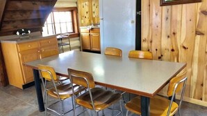 Uma área de refeições com uma mesa grande e quatro cadeiras, armários de madeira e uma janela com cortinas.