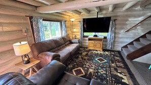 Interior de uma cabana de tronco com um sofá, um tapete, uma televisão e uma escada.