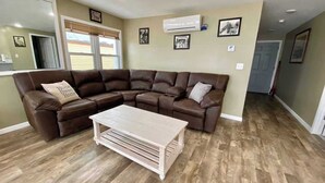 Ein Wohnzimmer mit einem braunen Ecksofa, einem hellen Holztisch und einer an der Wand montierten Klimaanlage.