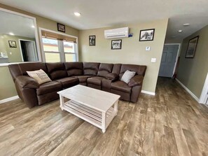 Ein Wohnzimmer mit einem braunen Ecksofa, einem hellen Holztisch und einer an der Wand montierten Klimaanlage.