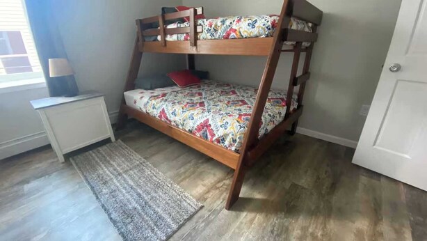 A bunk bed with a floral bedspread, a small white desk with a lamp, and a rug on a wooden floor. - Upstairs Apartment in Downtown Munising (Munising)