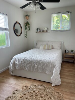 Ein Schlafzimmer mit einem Bett, einem Deckenventilator, einem Fenster mit Jalousien, einem Nachttisch mit einer Uhr und einem gewebten Teppich.
