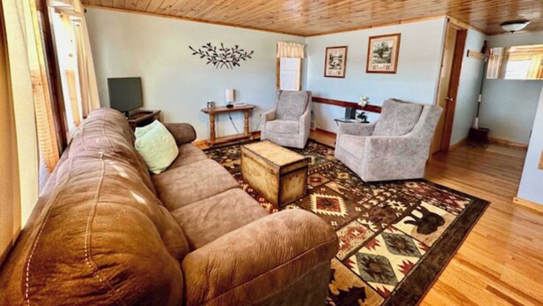 A cozy living room with a brown leather sofa, a small coffee table, and two armchairs. There is a patterned rug on the floor, a wall-mounted light fixture, and a television on a stand.