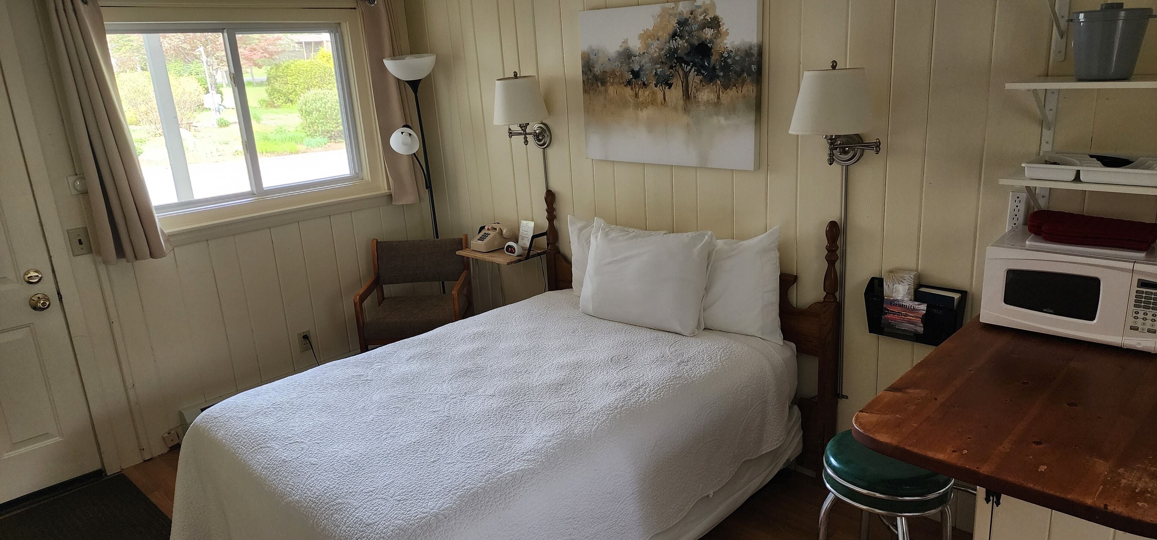 Cama bem arrumada em quarto com janela, quadro na parede, luminária, telefone e micro-ondas sobre uma mesa de madeira.