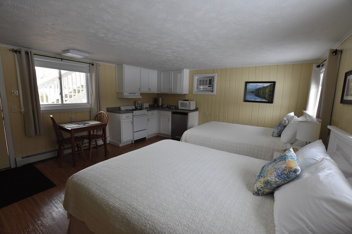 Una habitación con dos camas, una kitchenette pequeña, una mesa de comedor con sillas y una ventana con cortinas.