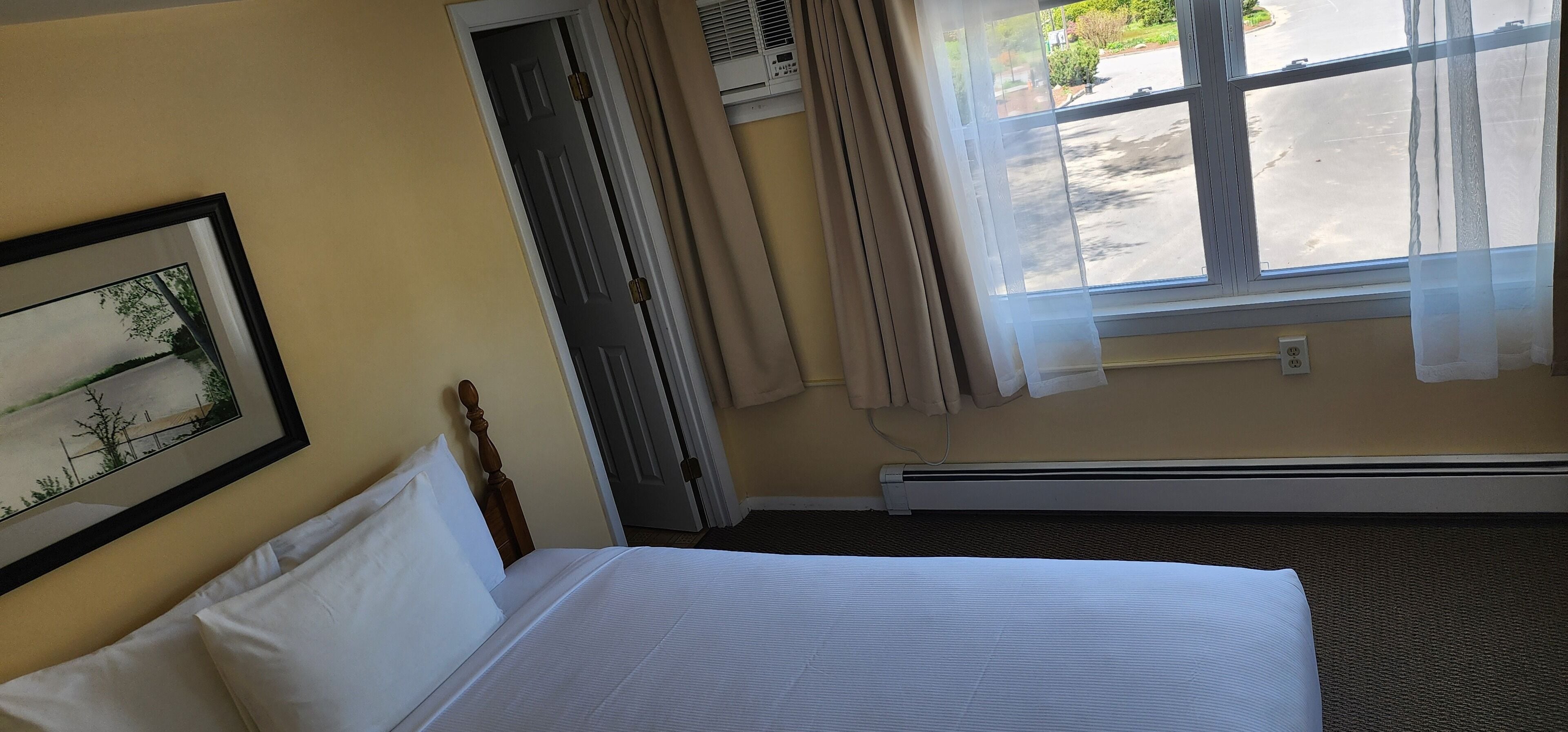 A hotel room with a bed, a framed picture on the wall, a window with curtains, and an air conditioning unit.