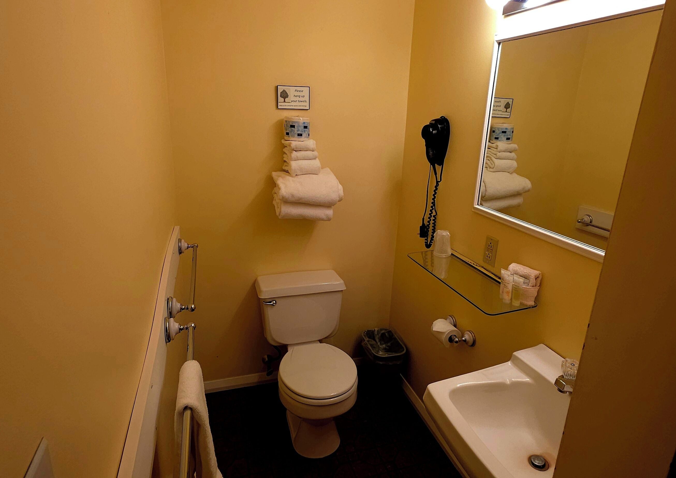 Et lille badeværelse med toilet, et glashylster med en hårtørrer, et håndklædestativ og et spejl.