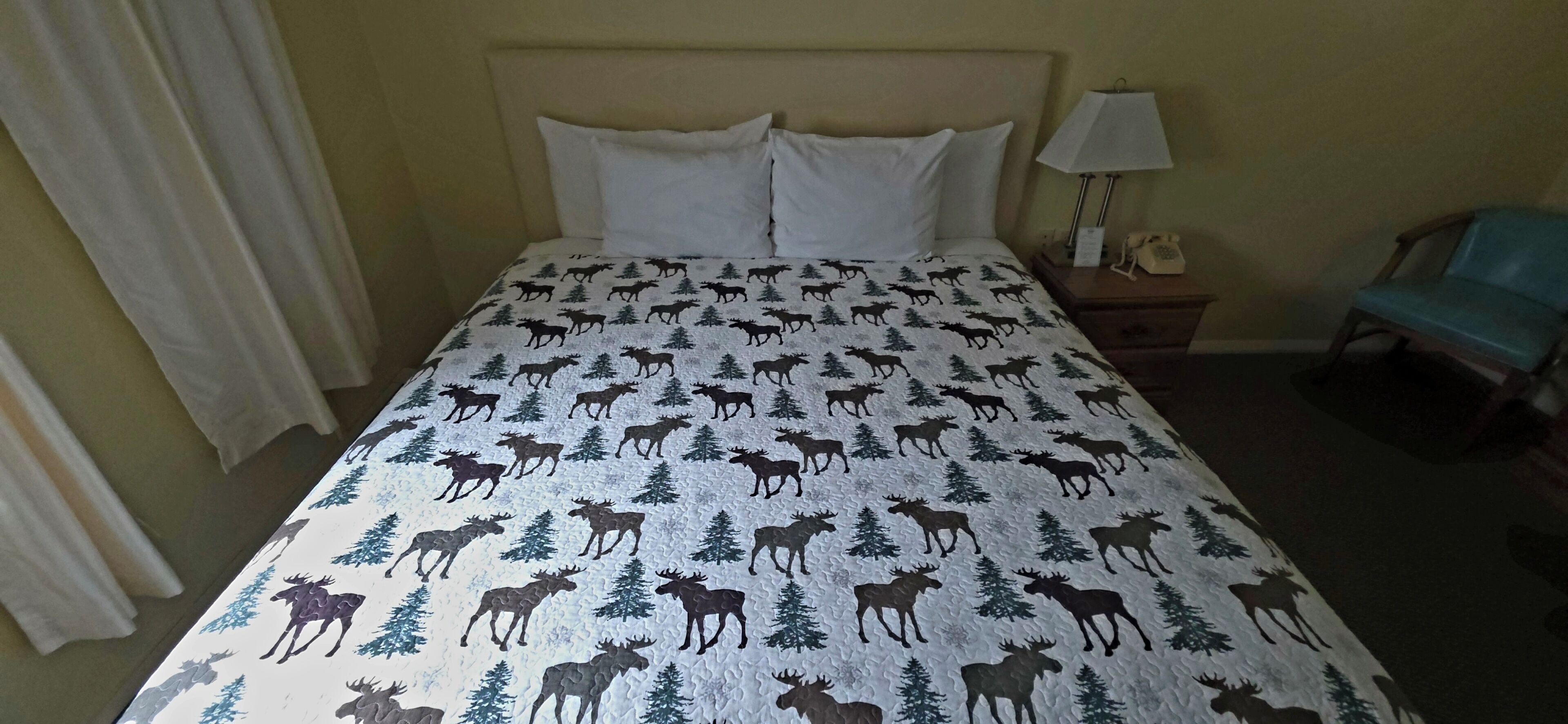 A bed with a moose and tree pattern, a bedside lamp, a chair, and a nightstand with a phone.
