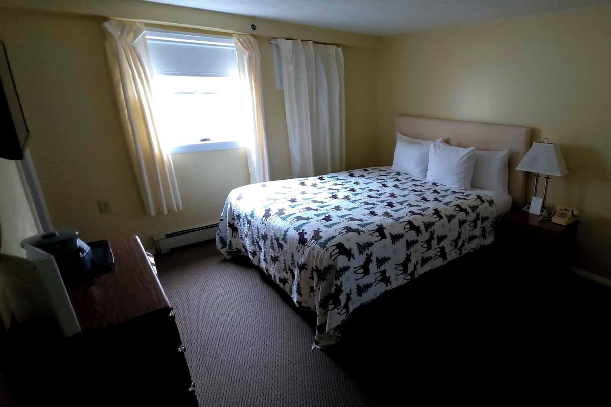 Ett hotellrum med en säng, ett nattduksbord, en lampa, ett fönster med gardiner och ett skrivbord med en stol.