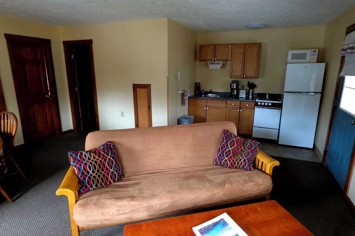 A living room with a sofa, two chairs, a coffee table, and a kitchen area with cabinets, a stove, and a refrigerator.