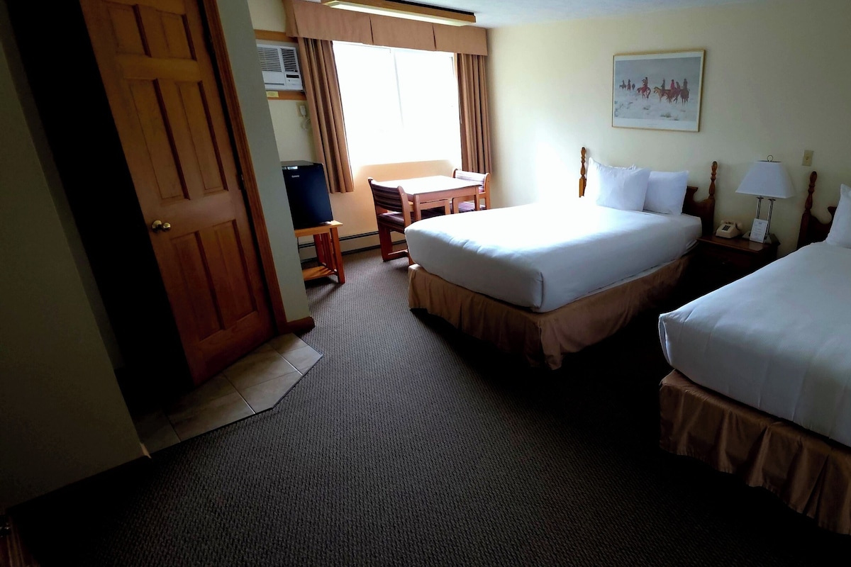 A hotel room with two beds, a desk, a chair, a lamp, and two paintings on the walls.