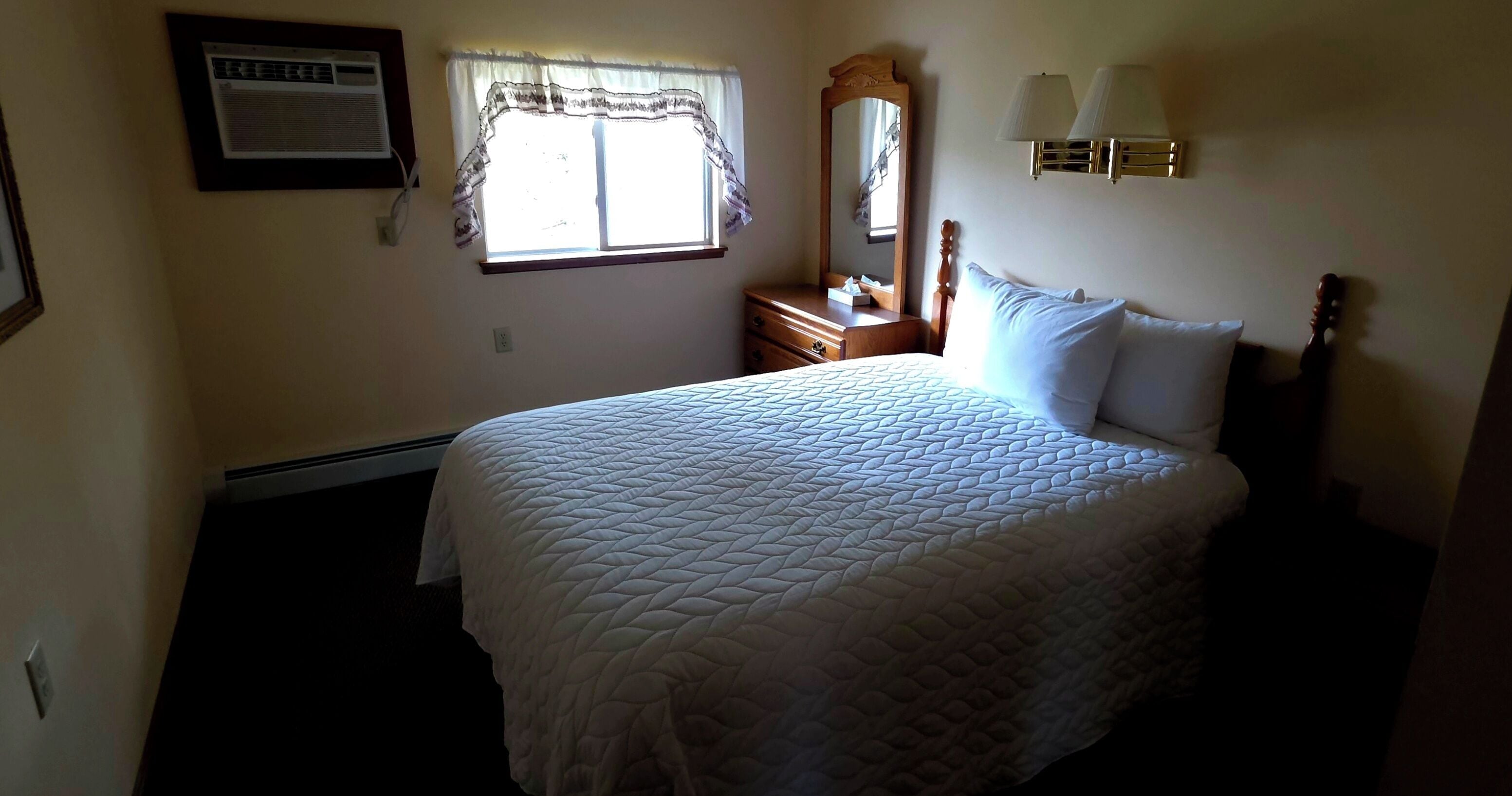 A bedroom with a bed, a window with curtains, a dresser, a mirror, and wall-mounted lamps.