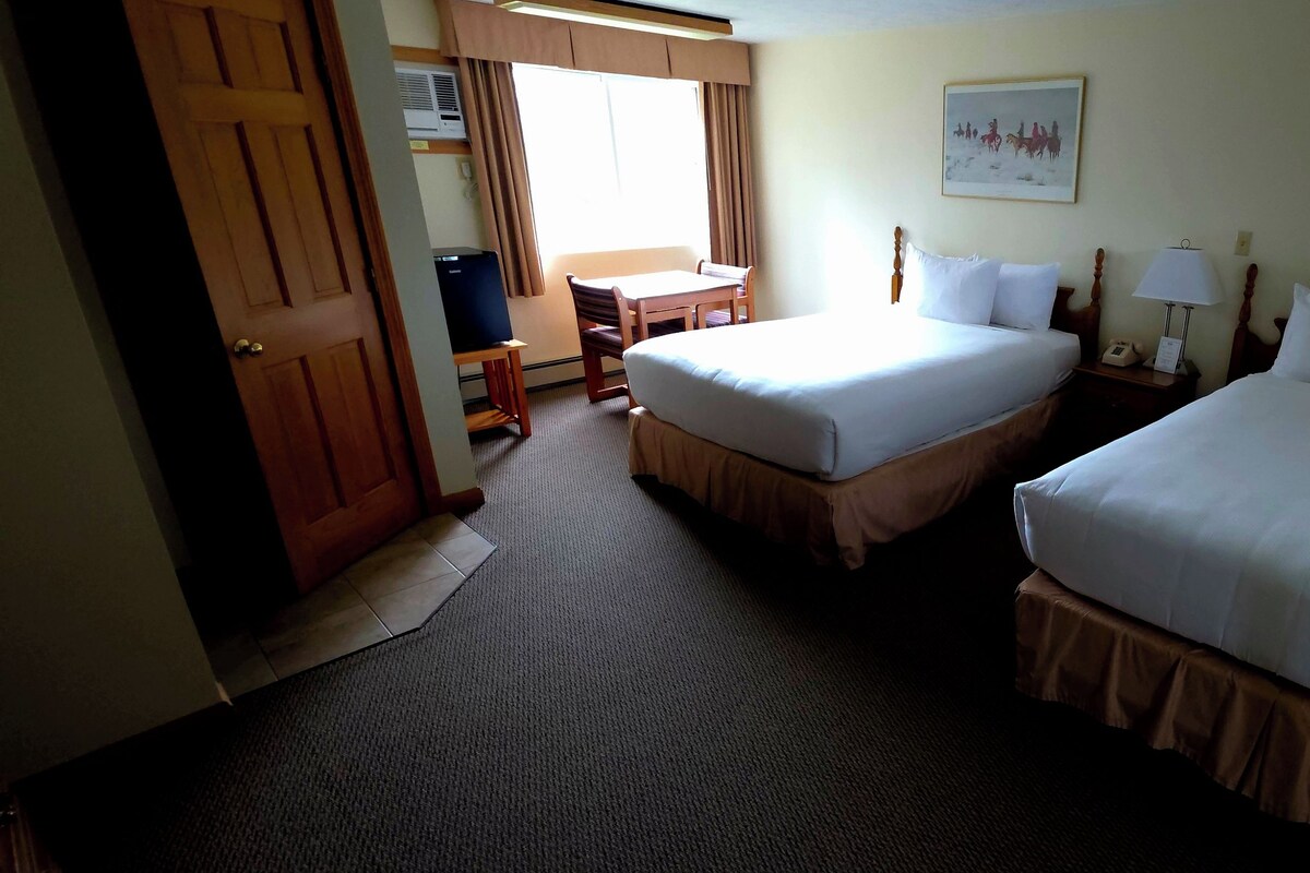 Habitación de hotel con dos camas, un escritorio, una silla, una lámpara y dos cuadros en las paredes.