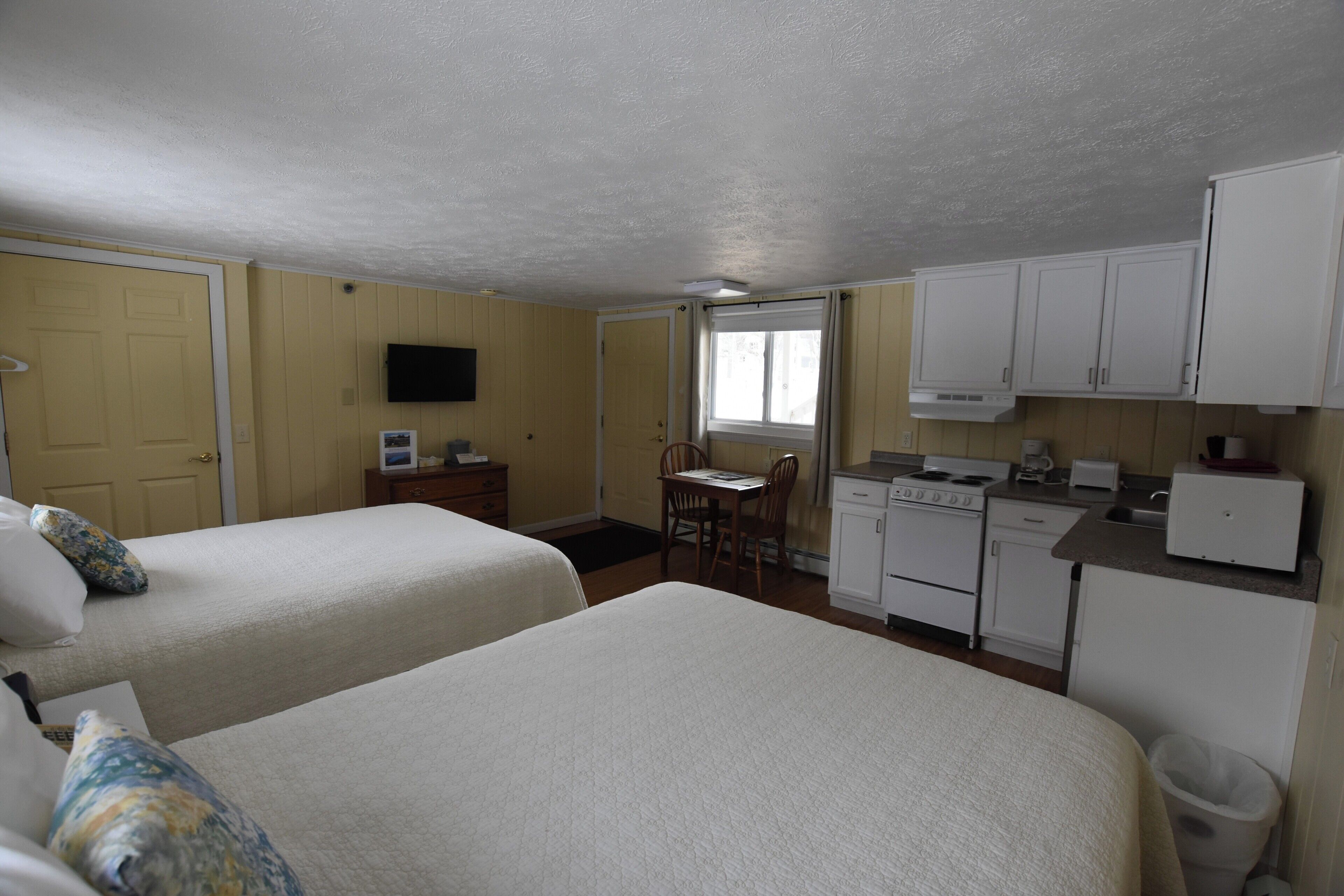 A room with two beds, a small kitchenette, a dining table with chairs, and a television mounted on the wall.
