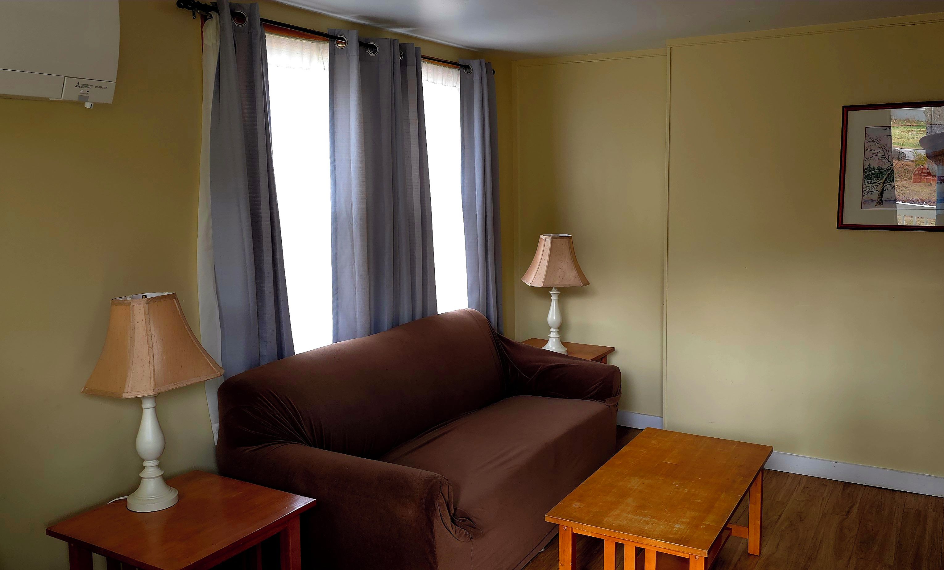A room with a brown sofa, two small tables with lamps, a window with curtains, and a framed picture on the wall.