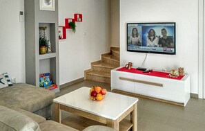 A modern living room with a sofa, a coffee table with fruit, a TV displaying a news program, a staircase, and a red shelf with plants. - Lovely home in Vodnjan (Vodnjan)