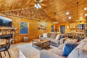 A cozy log cabin interior with a fireplace, wooden walls, a sofa, a coffee table, and a dining area. - New Listing! Family Gameroom Cabin+Fire Pit+Hot Tub+Dog Friendly+Close to BR! (Morganton)