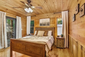 A wooden cabin bedroom with a bed, a nightstand, a ceiling fan, and a window with curtains. - New Listing! Family Gameroom Cabin+Fire Pit+Hot Tub+Dog Friendly+Close to BR! (Morganton)