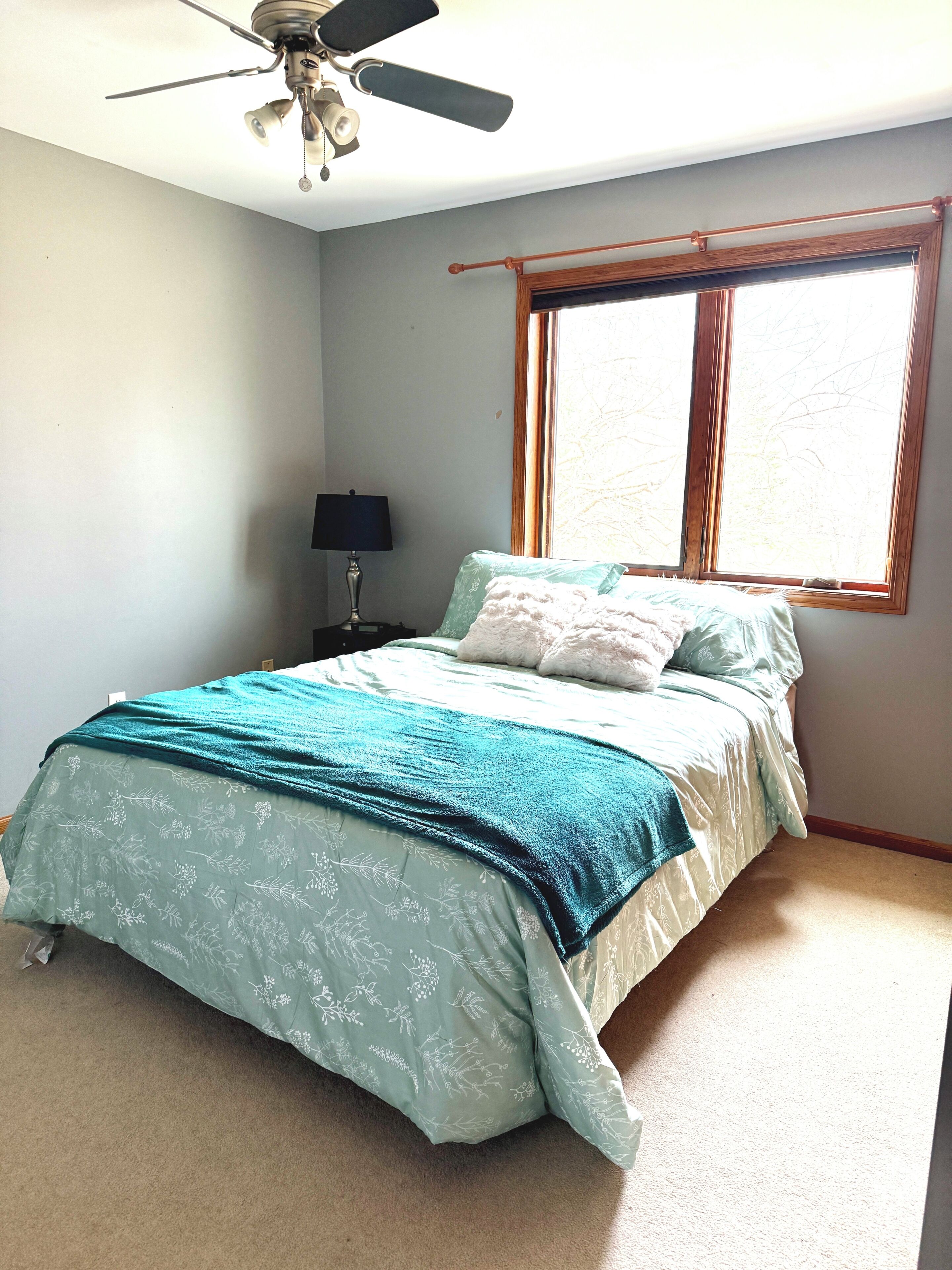 A bedroom with a bed, a ceiling fan, a nightstand, a lamp, and a window.