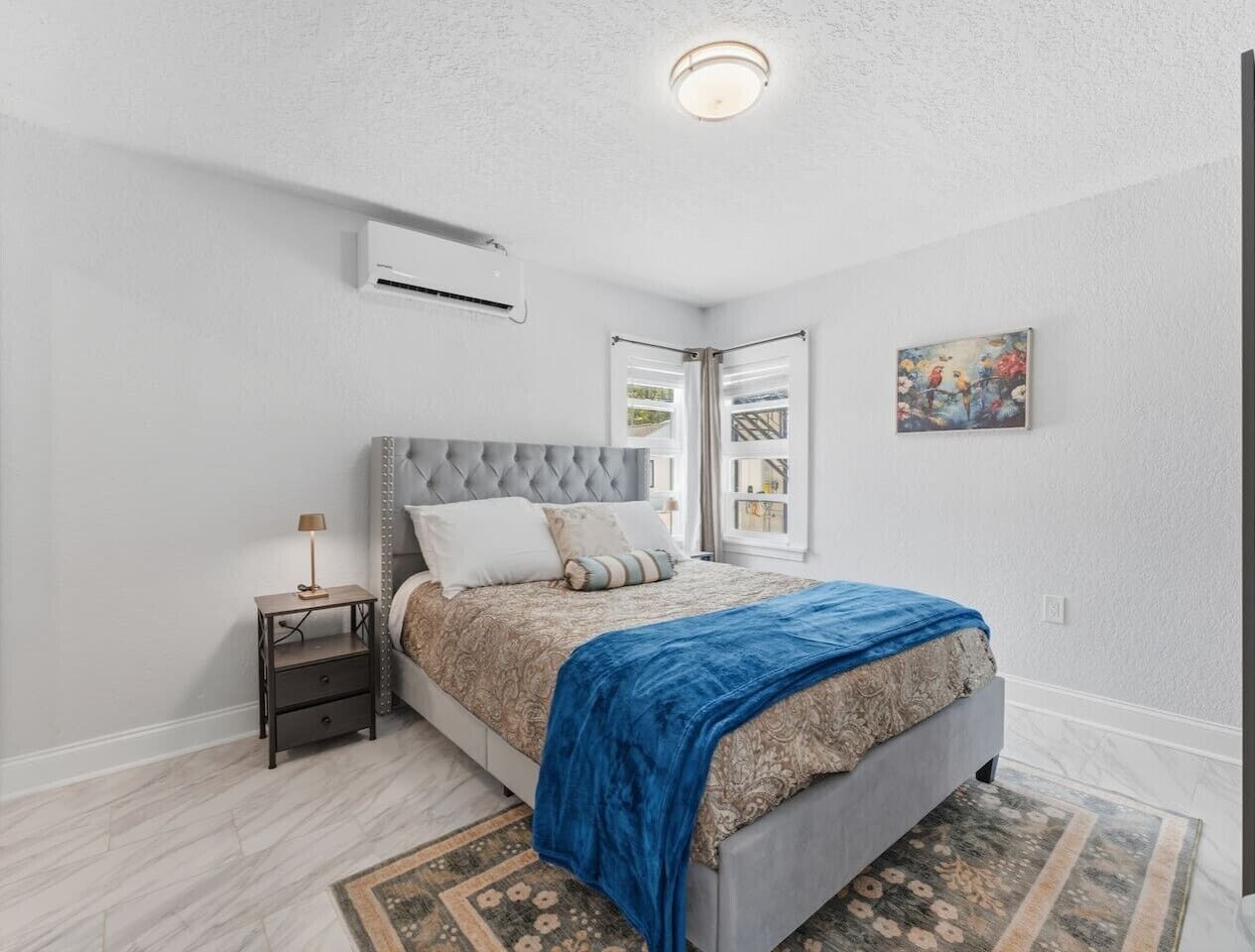 Un dormitorio con cama, mesita de noche, lámpara, equipo de aire acondicionado y un cuadro en la pared.