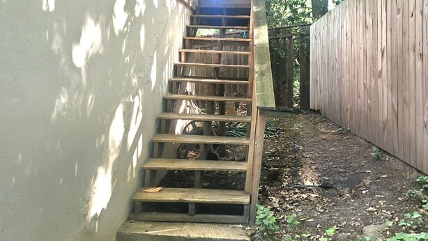 Een houten trap die leidt naar een balkon, omgeven door een houten hek en groen.