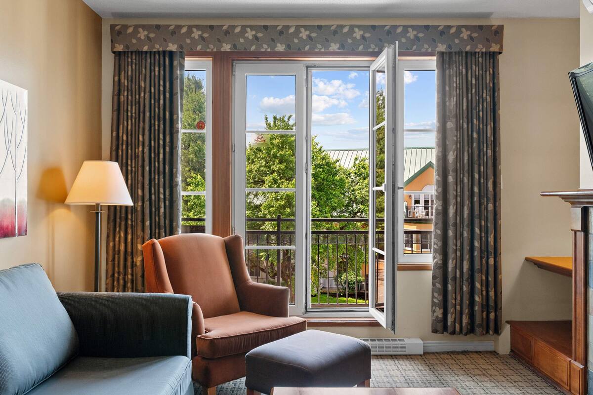 Habitación con sofá, sillón, reposapiés, una lámpara y vista a un balcón exterior por medio de una puerta de vidrio.