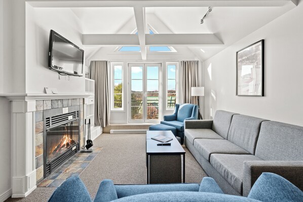 A modern living room with a fireplace, a flat-screen TV, a sofa, a coffee table, and large windows.