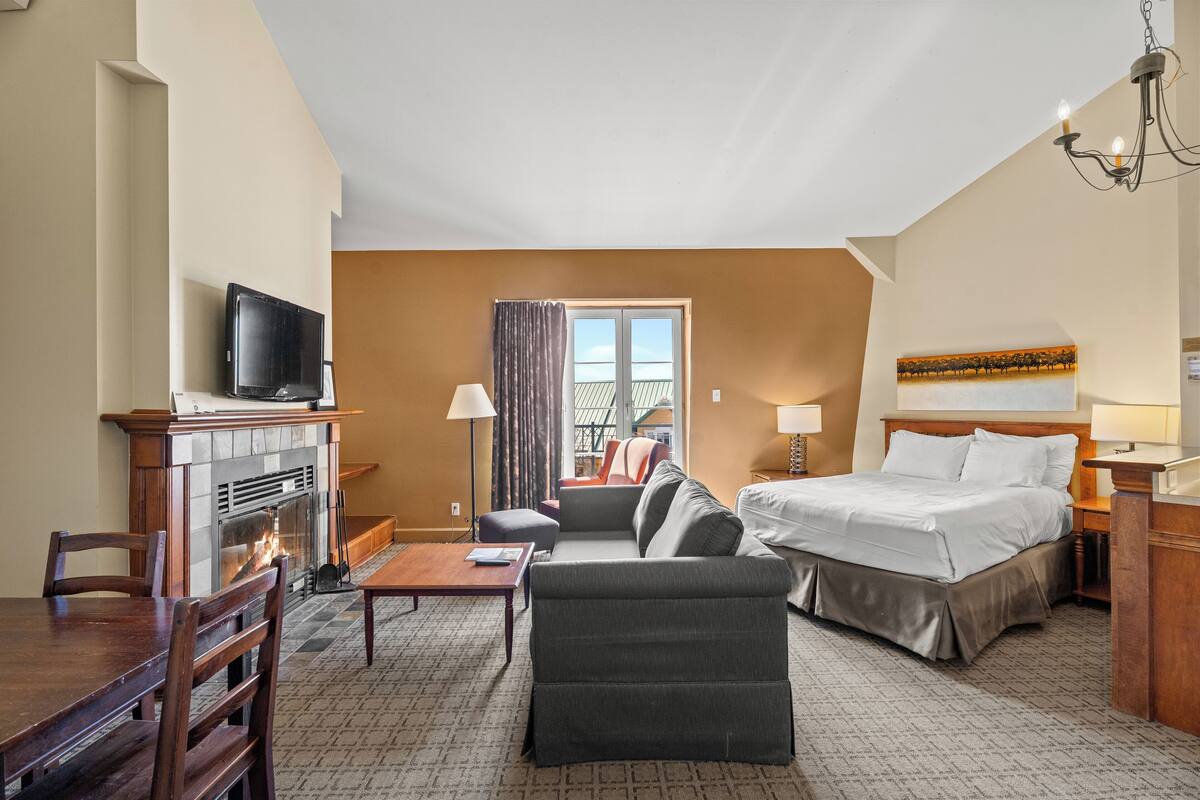 Habitación de hotel con cama, sofá, televisión, chimenea, mesa de comedor y ventana con cortinas.