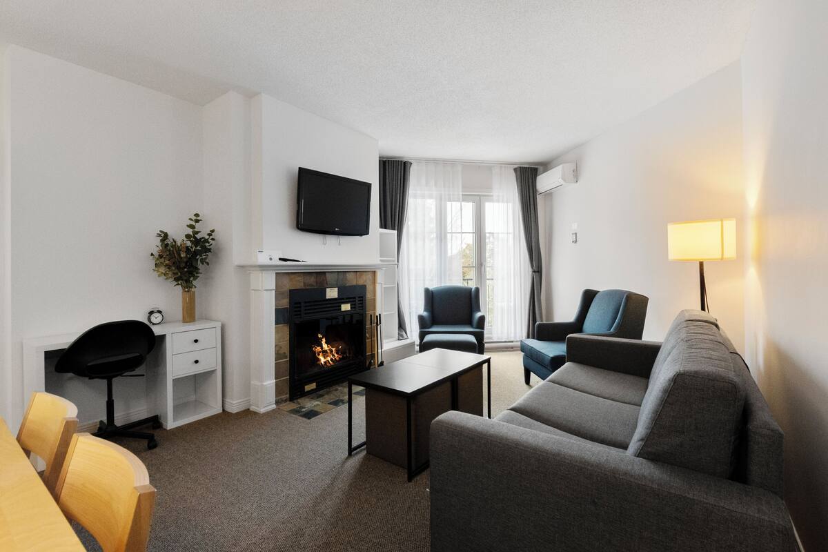 Habitación de hotel con chimenea, un televisor de pantalla plana colocado encima, un sofá, un escritorio con una silla y una lámpara.