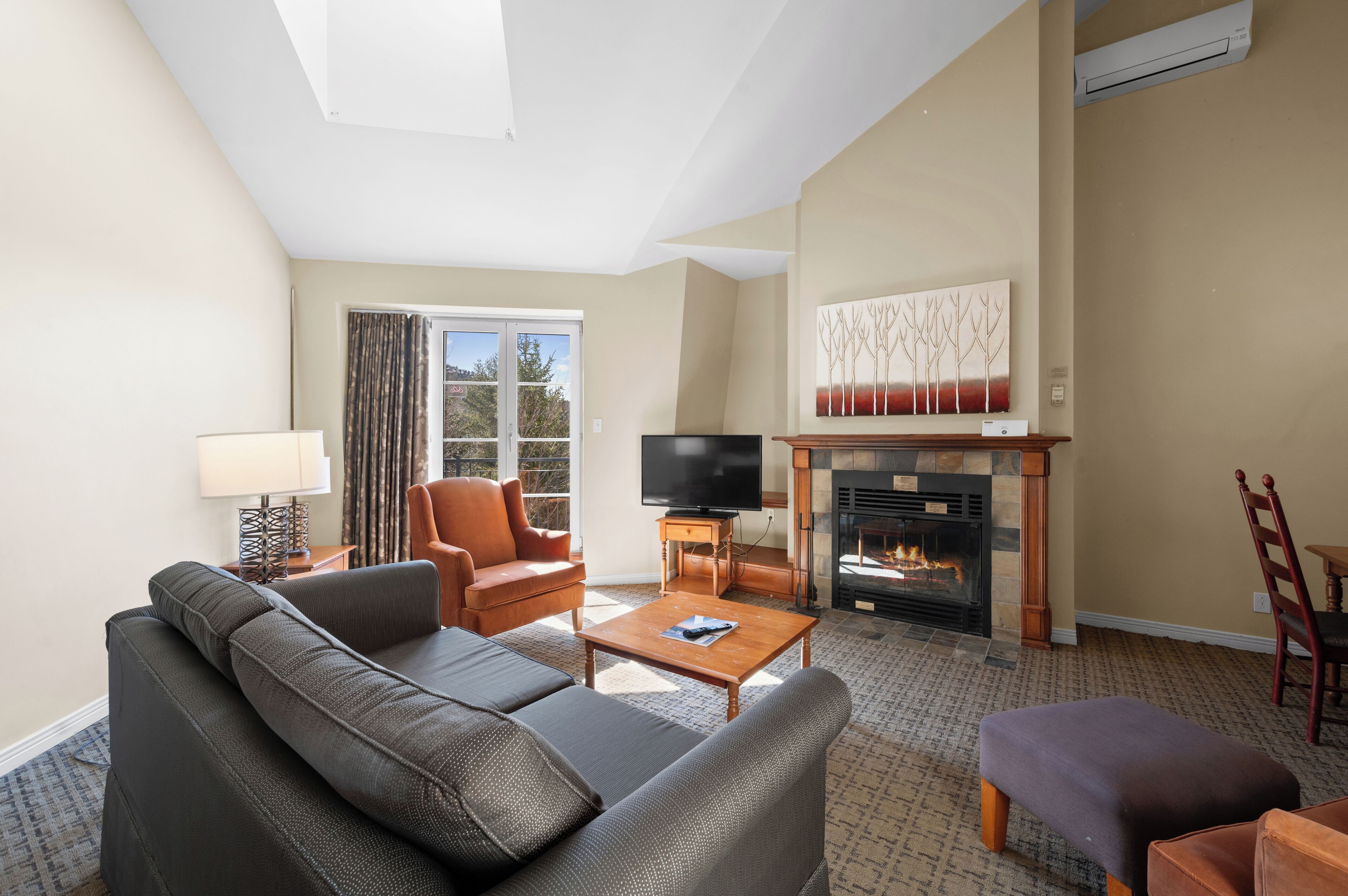 Ein Wohnzimmer mit Sofa, Sesseln, Couchtisch, Kamin, Fernseher und Blick durch ein Fenster.