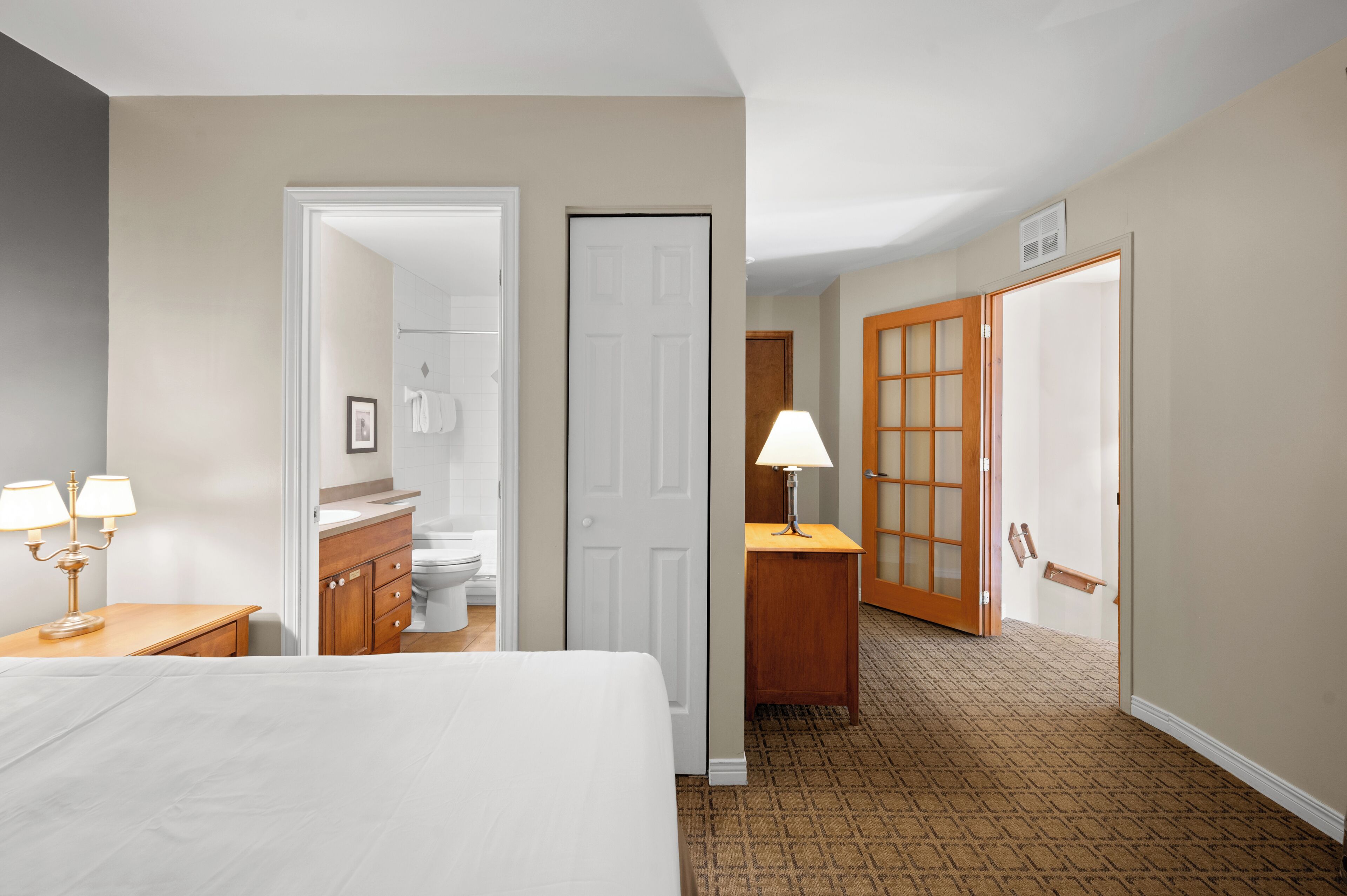 A hotel room with a bed, bedside table, lamp, wooden dresser, and a bathroom visible through an open door.
