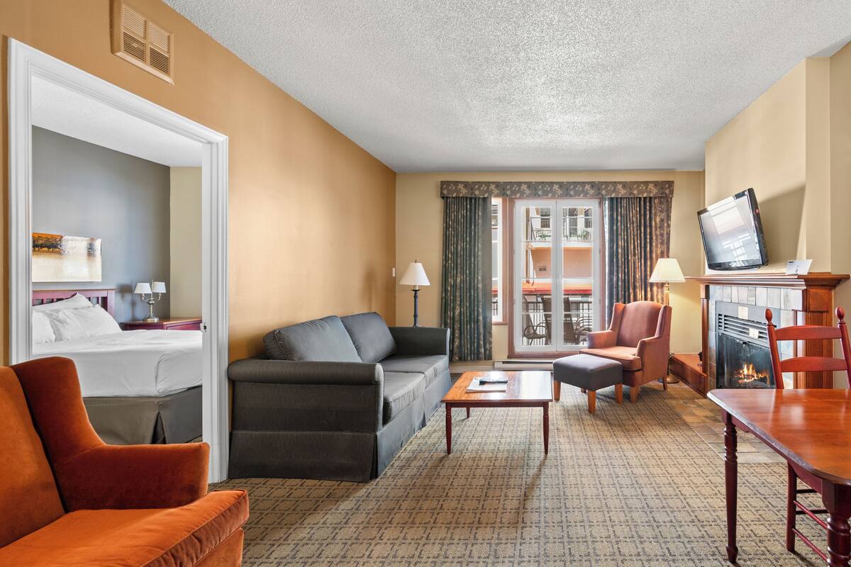 Habitación de hotel con cama, sofá, mesa de centro, televisor, chimenea y balcón con cortinas.