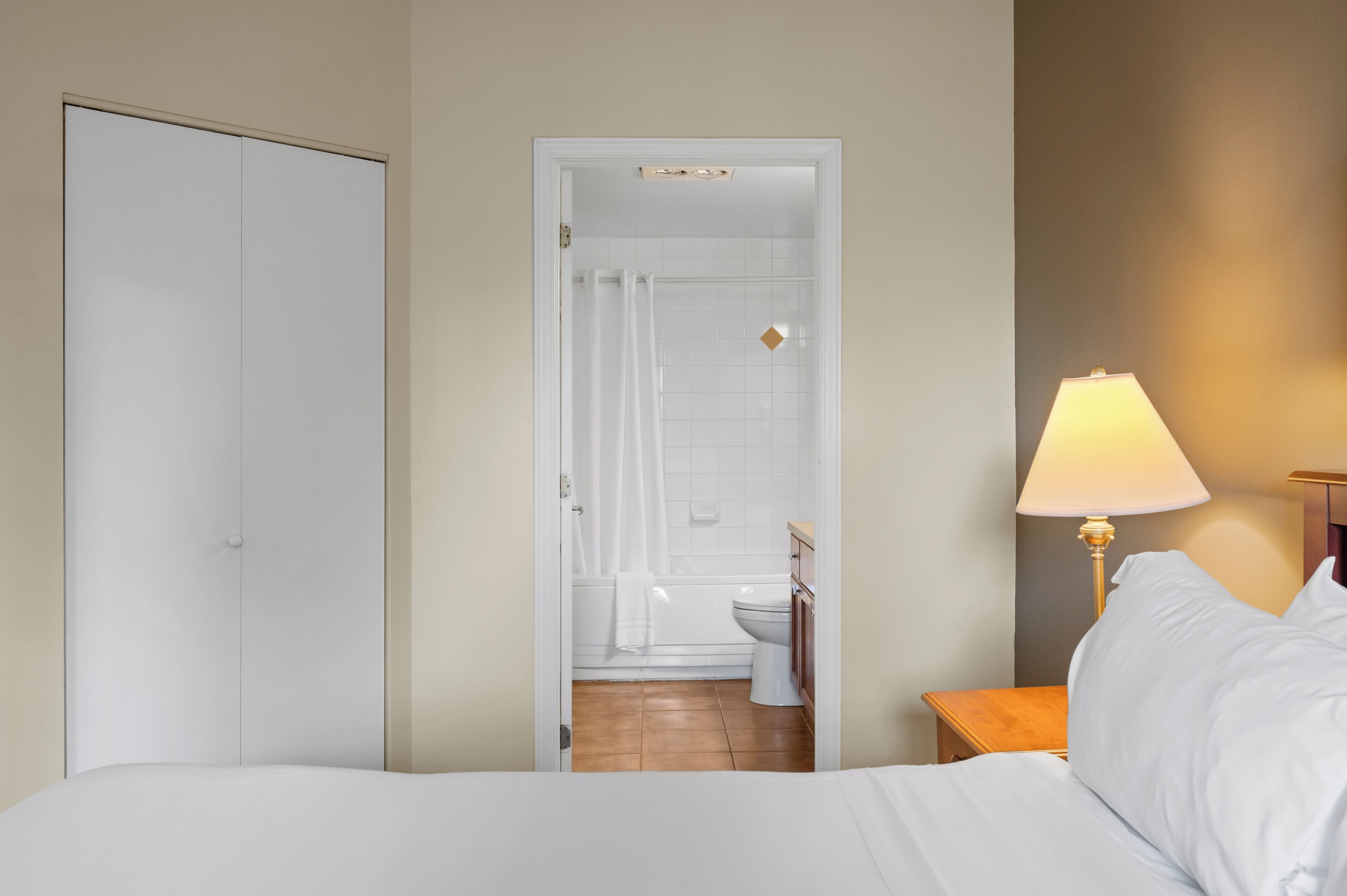 A bedroom with a bed, a nightstand, a lamp, and an open door leading to a bathroom with a toilet and shower.