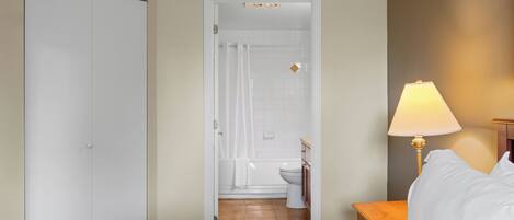 A bedroom with a bed, a nightstand, a lamp, and an open door leading to a bathroom with a toilet and shower.