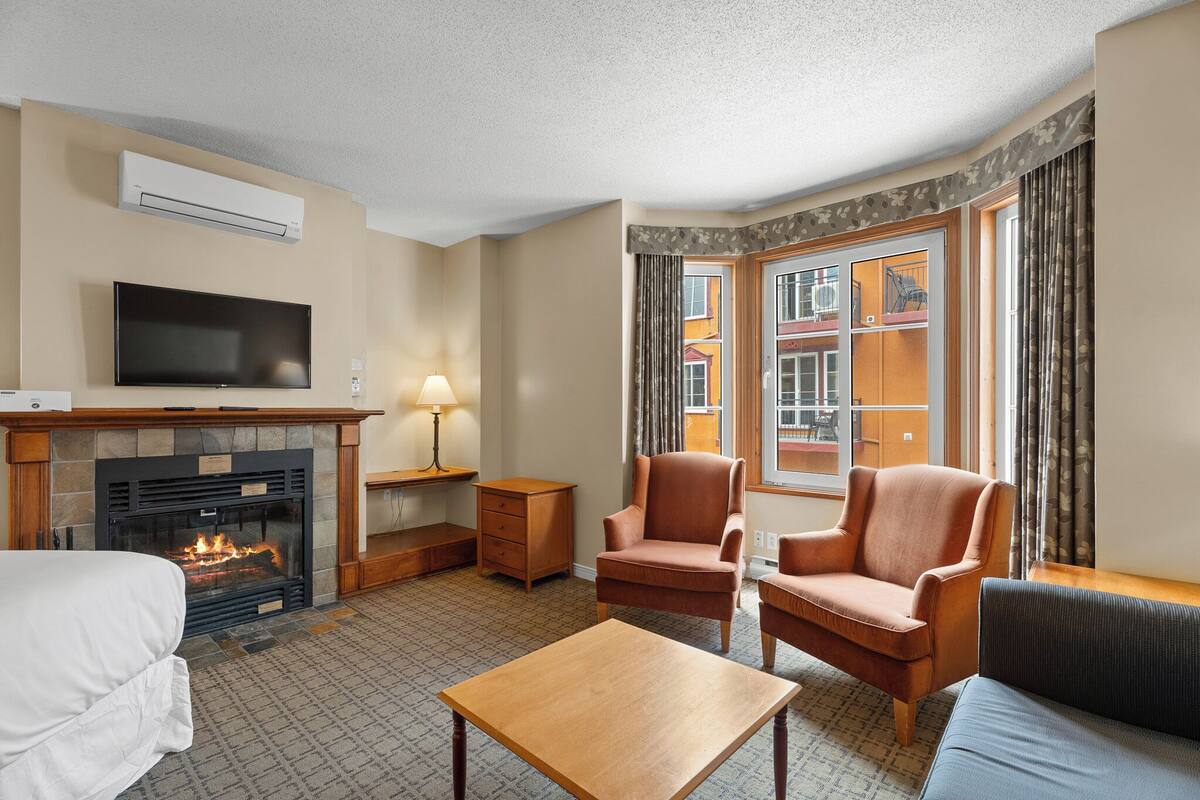 Habitación de hotel con chimenea, televisor, sofá, sillón, mesa y ventana con vistas a edificios.