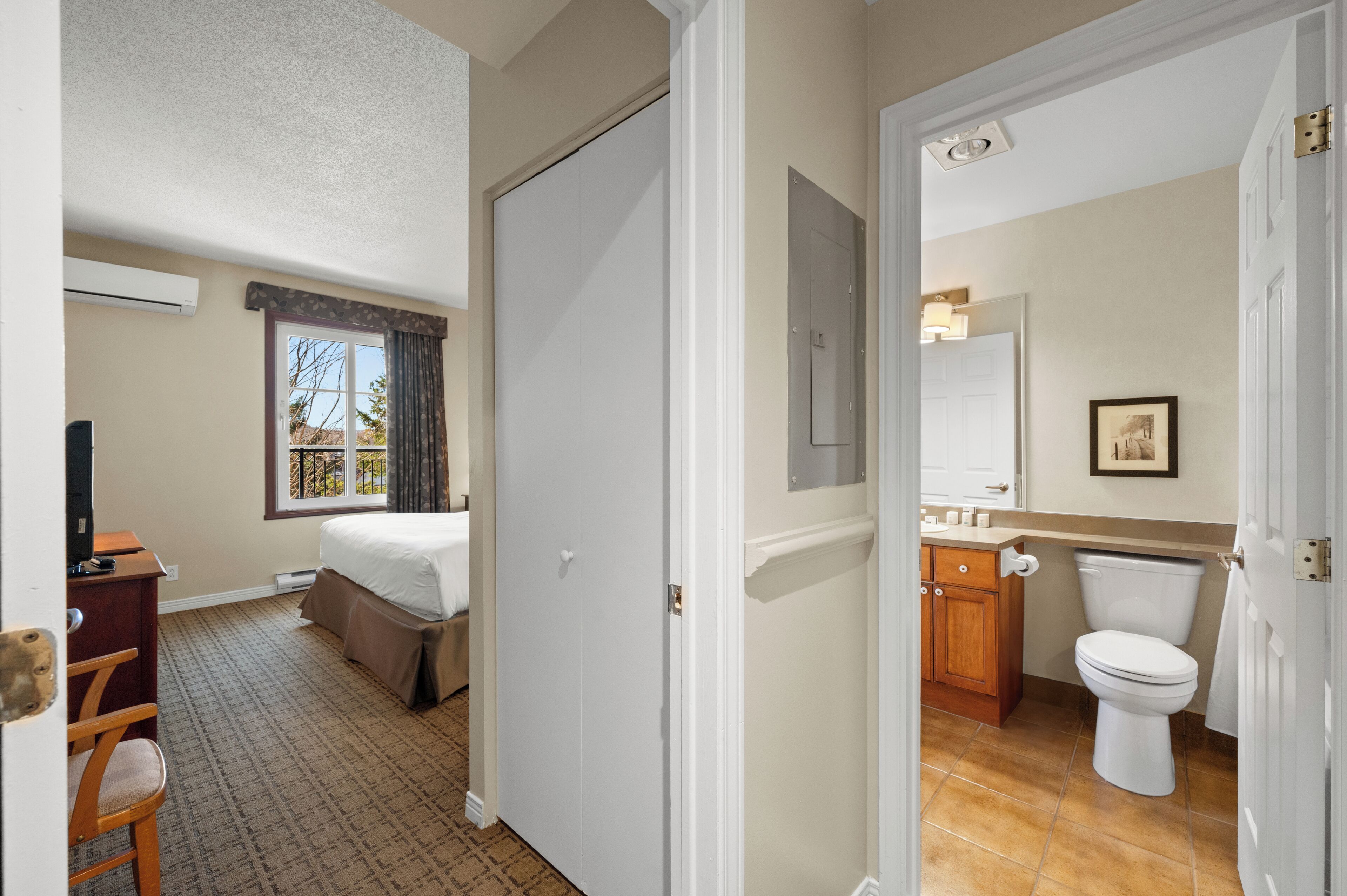Hotelkamer met een bed, bureau, stoel, raam met gordijnen, en een badkamer met toilet, wastafel en spiegel.