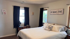 Une chambre à coucher avec un lit, deux fenêtres avec des rideaux, une table de chevet avec un ventilateur, un panneau indiquant une zone où il est interdit de faire du bruit et une petite plante.