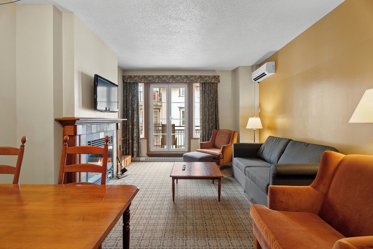 Une chambre d’hôtel avec une table à manger en bois, un canapé, une chaise, un téléviseur, une cheminée et un balcon avec des rideaux.