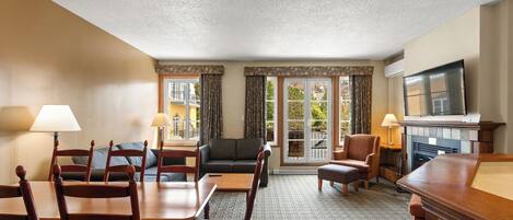 Habitación de hotel con mesa de comedor, sillas, sofá, chimenea, televisor y ventanas amplias con cortinas.