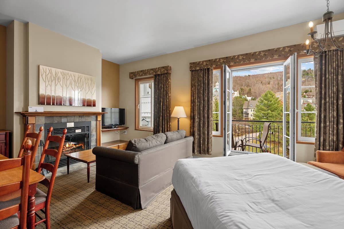A hotel room with a fireplace, sofa, bed, dining table, chairs, and a balcony with a view.