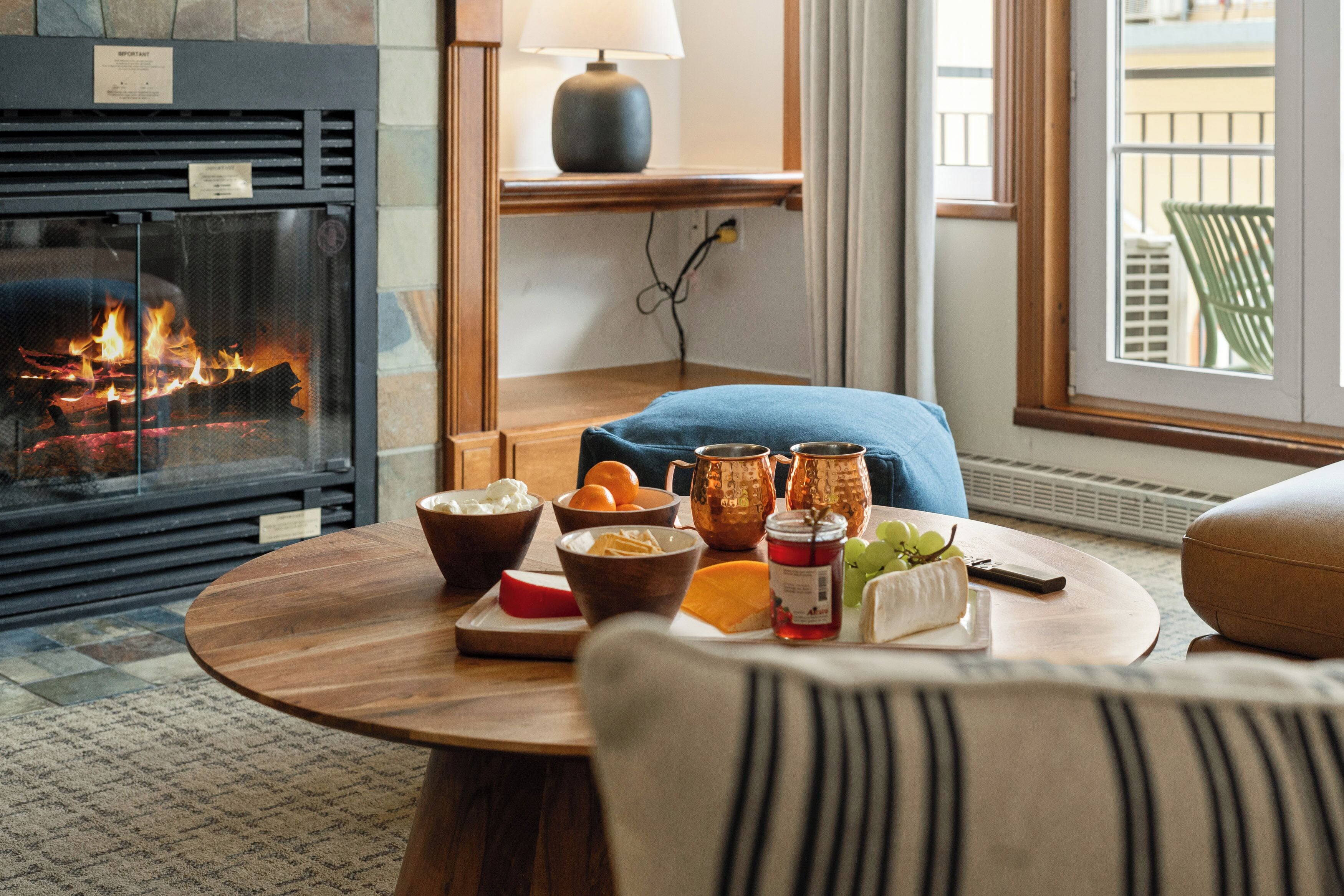 A cozy room with a fireplace, a round wooden table with food and drinks, a lamp, and a view of the outdoors.