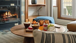 A cozy room with a fireplace, a round wooden table with food and drinks, a lamp, and a view of the outdoors. - Le Deslauriers by RVMT (Mont-Tremblant)