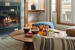 Ein gemütliches Zimmer mit Kamin, einem runden Holztisch mit Speisen und Getränken, einer Lampe und Blick ins Freie.