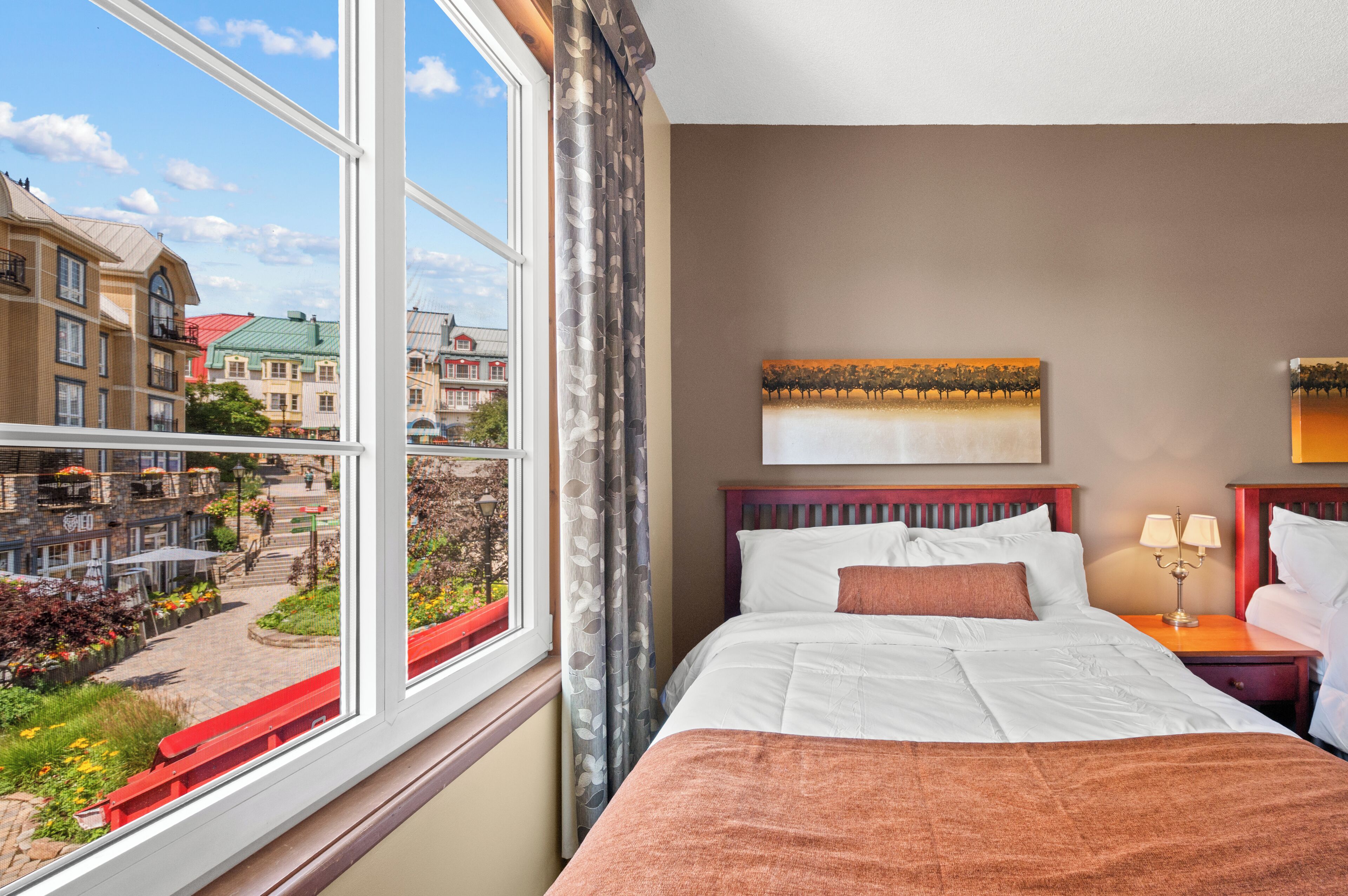 A hotel room with a large window, a bed with white linens, a headboard, a nightstand with a lamp, and a painting on the wall.