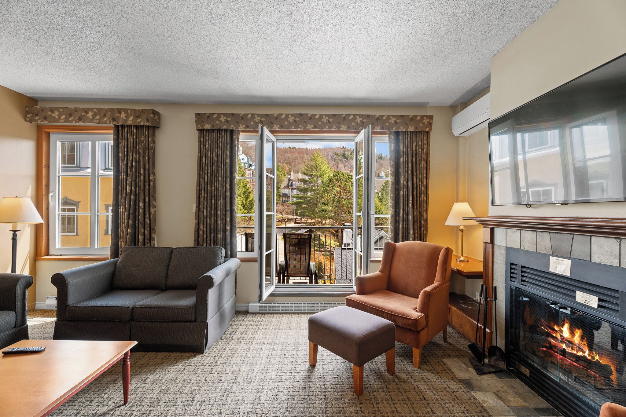 Ein Hotelzimmer mit einer Couch, einem Sessel, einem Hocker, einem Kamin und einem Balkon mit Blick.