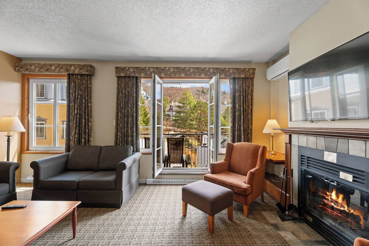 A hotel room with a sofa, armchair, ottoman, fireplace, and a balcony with a view.