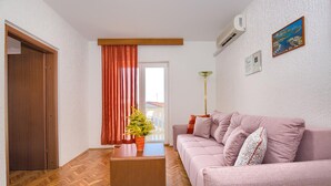 Una sala de estar con piso de madera, un sofá, una mesa de centro, una planta en maceta, una ventana con cortinas y un equipo de aire acondicionado.