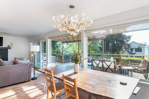 A spacious living area with a large wooden table, a sofa, and a dining area with outdoor seating. - Water and Mountain Views, 2 Kitchens, 2 Decks, Sunny Hilltop Retreat, Sleeps 11 (Seattle)