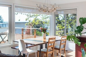 A dining area with a wooden table and chairs, a vase with flowers, and a view of the outdoors through large windows. - Water and Mountain Views, 2 Kitchens, 2 Decks, Sunny Hilltop Retreat, Sleeps 11 (Seattle)