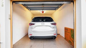 A white Mazda CX-5 parked inside a garage with a closed garage door, a visible fire alarm, and a LHAUL construction sign on the right. - 2BR Townhome, Private, Monthly Stays Welcome! (Land O' Lakes)